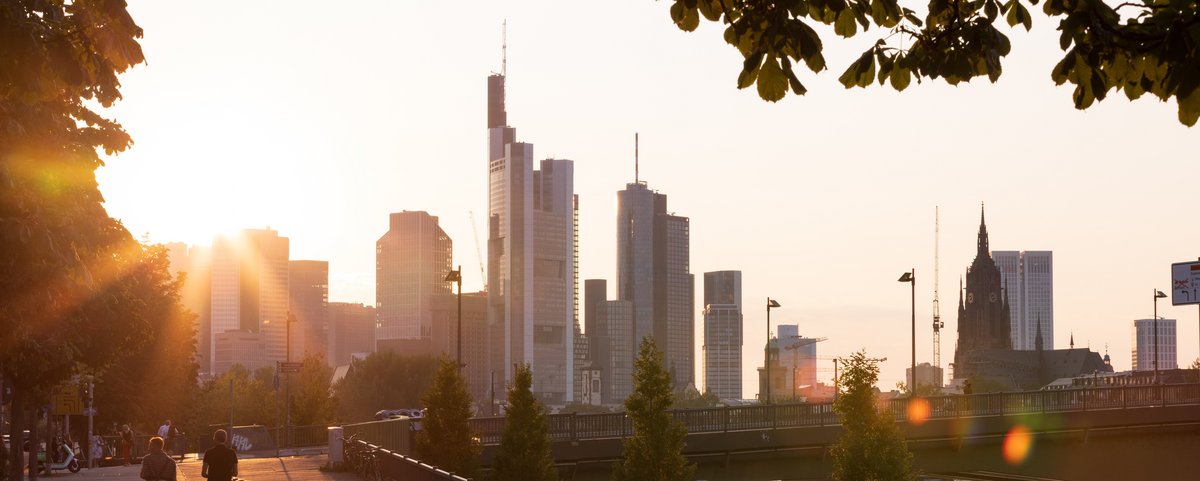 Sonnenuntergang über der Frankfurter Skyline mit Hochhäusern, Brücke und Menschen, die am Flussufer spazieren und joggen