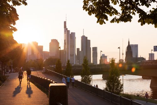 Sonnenuntergang über der Frankfurter Skyline mit Hochhäusern, Brücke und Menschen, die am Flussufer spazieren und joggen