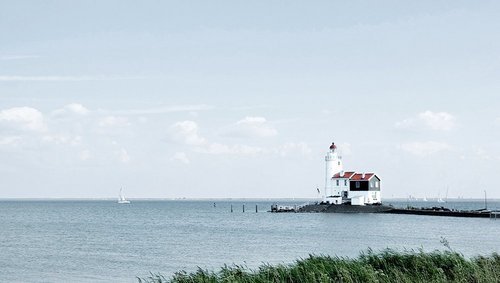 Ein Leuchtturmhaus am Ijsselmeer mit Schilf im Vordergrund und Segelbooten auf dem Meer, unter einem bewölkten Himmel.