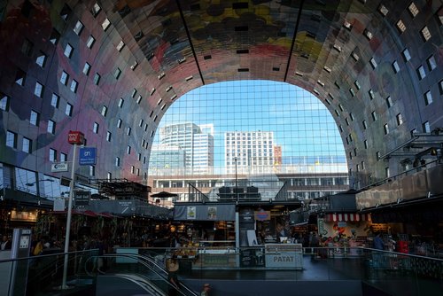 Innenansicht der Markthalle in Rotterdam mit bunten Wandfliesen, zahlreichen Verkaufsständen und Blick auf moderne Gebäude durch das große Fenster.