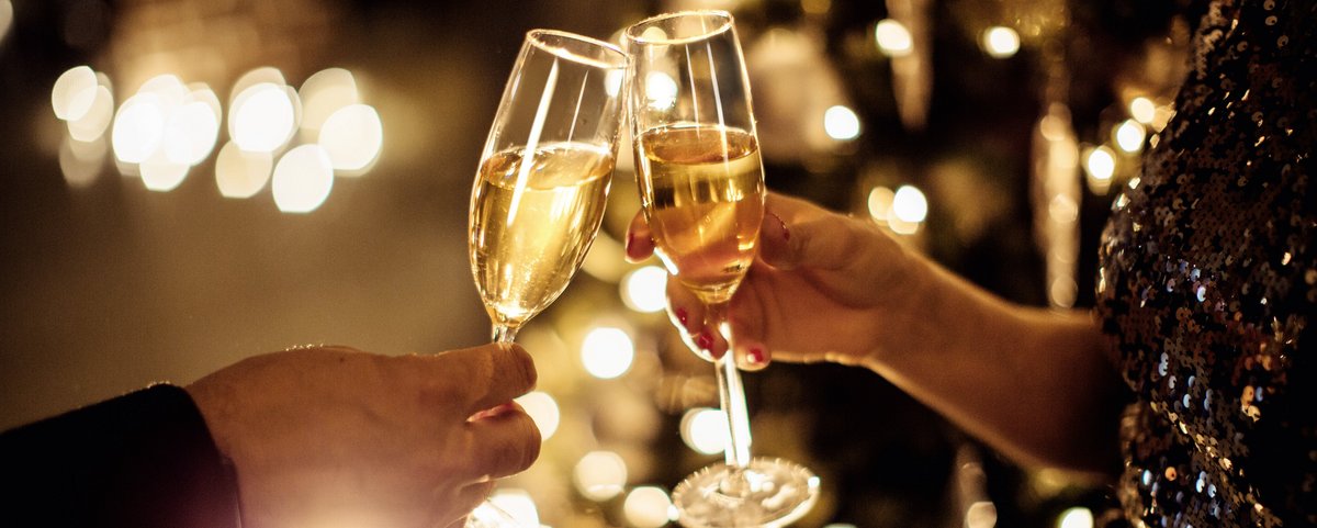 Zwei Hände stoßen mit gefüllten Sektgläsern vor einem festlich beleuchteten Hintergrund an.