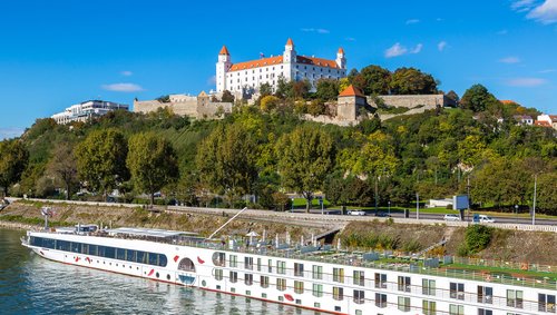 Blick auf die Bratislavaer Burg auf einem bewaldeten Hügel mit einem weißen A-ROSA Flusskreuzfahrtschiff auf der Donau im Vordergrund