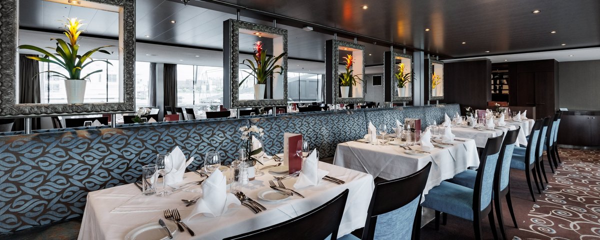 Stilvoll eingedeckter Restaurantbereich der A-ROSA CLEA mit Fenstern, Blumendeko und gedämpfter Beleuchtung.