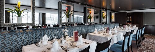 Stilvoll eingedeckter Restaurantbereich der A-ROSA CLEA mit Fenstern, Blumendeko und gedämpfter Beleuchtung.