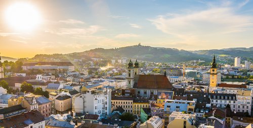 Panoramablick über die Linz bei Sonnenuntergang. Mit den Dächer und Häuser der Altstadt, den Türmen der Kirche und dem Uhrturm des Alten Rathause. Im Hintergrund bewaldete Hügel, der Himmel ist blau mit leichten Wolken. 