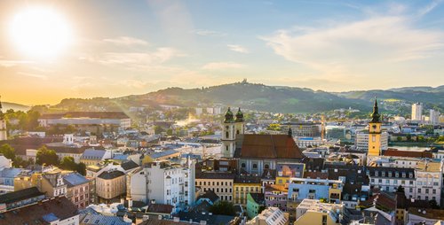 Panoramablick über die Linz bei Sonnenuntergang. Mit den Dächer und Häuser der Altstadt, den Türmen der Kirche und dem Uhrturm des Alten Rathause. Im Hintergrund bewaldete Hügel, der Himmel ist blau mit leichten Wolken. 
