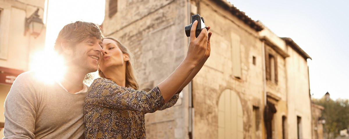 Ein Paar sitz in der Altstadt von Arles auf einer Mauer und macht mit einer Digitalkamera ein Bild von sich. Im Hintergrund historische Gebäude und die untergehende Sonne.