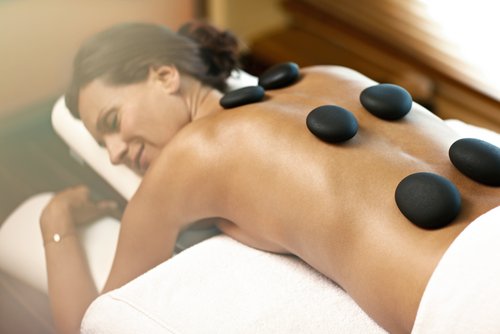 Eine Frau liegt entspannt und lächelnd auf einer Massageliege, während heiße Steine auf ihrem Rücken platziert sind, umgeben von einem ruhigen Spa-Ambiente.