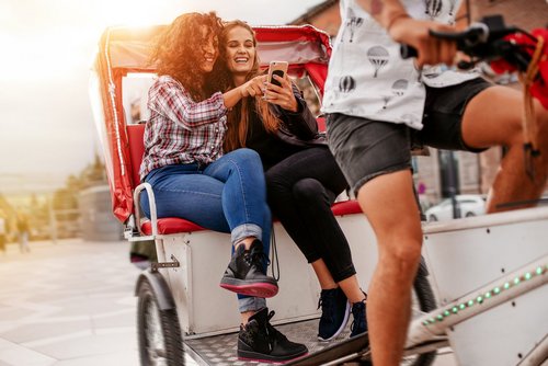 Zwei Personen lassen sich auf einer Rikscha durch eine belebte Stadtstraße fahren und schauen auf ihr Handy.