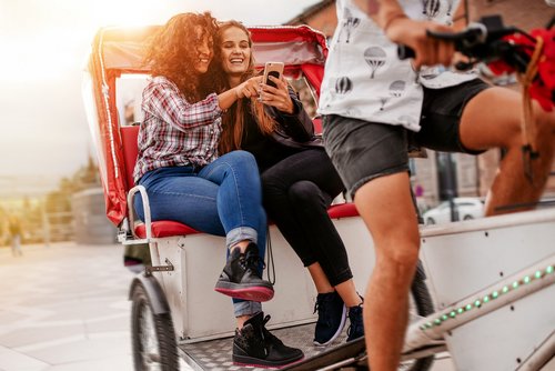 Zwei Personen lassen sich auf einer Rikscha durch eine belebte Stadtstraße fahren und schauen auf ihr Handy.