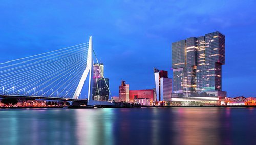 Skyline von Rotterdam bei Abenddämmerung mit der Maas im Vordergrund, mit modernen Hochhäusern und der Erasmusbrücke. Die Lichter der Stadt reflektieren im Wasser.