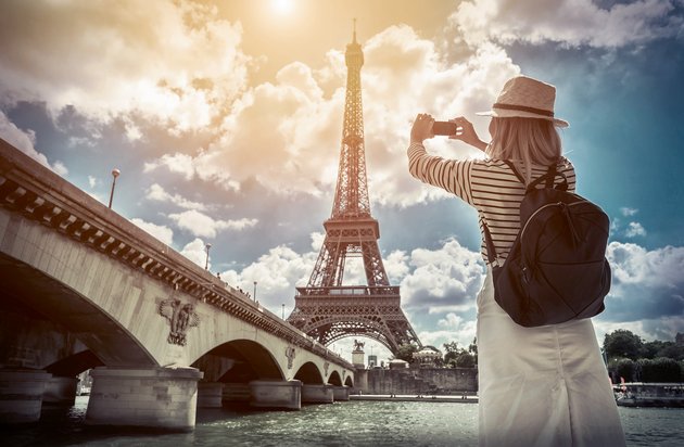 Touristin mit Strohhut und Rucksack fotografiert den Eiffelturm bei sonnigem Himmel von der Seine-Brücke in Paris.