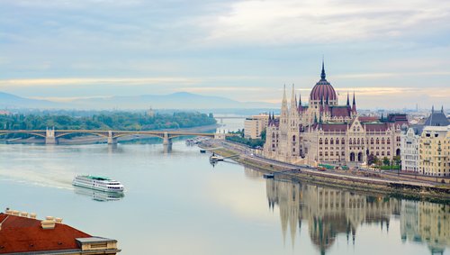 Blick auf das Parlamentsgebäude von Budapest am Flussufer. Das Gebäude spiegelt sich in der Donau und ein Flussschiff ist im Vordergrund.