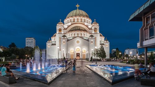Der beleuchtete Dom des Heiligen Sava in Belgrad bei Nacht. Im Vordergrund schmücken Springbrunnen und Laternen den Eingang und viele Menschen sitzen auf dem Vorplatz