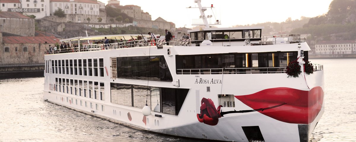 Flusskreuzfahrtschiff A-ROSA ALVA auf dem Douro vor einer Stadt mit historischen Gebäuden und roten Dächern.