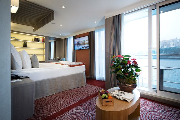 Eine stilvoll eingerichtete Suite auf der A-ROSA ALVA mit Doppelbett, Sofa, Schreibtisch und bodentiefen Fenstern und Balkon bei Tageslicht.