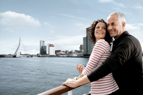 Ein Paar lehnt an der Reling eines Flusskreuzfahrtschiffs mit Blick auf den Rhein und die Skyline von Rotterdam.
