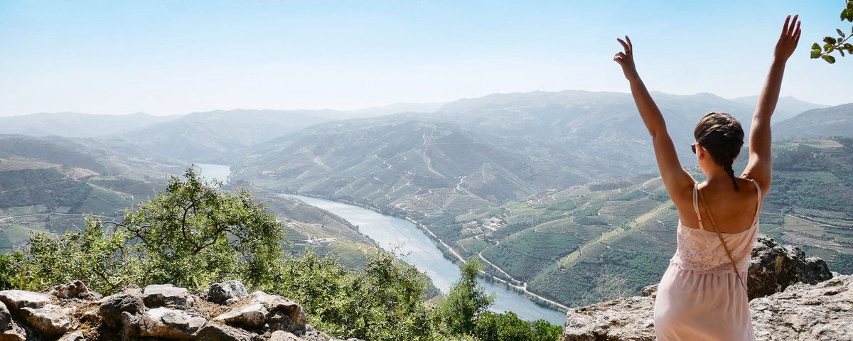 Frau in weißem Kleid steht auf Felsen und hebt die Arme über einen Fluss und grüne Hügel im Tal.