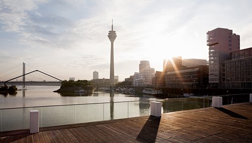 Blick von einer Holzbrücke mit Glasgeländer auf Düsseldorf mit dem Rheinturm und modernen Gebäuden am Flussufer, bei Sonnenschein und dem reflektierenden Rhein mit Brücken im Hintergrund.