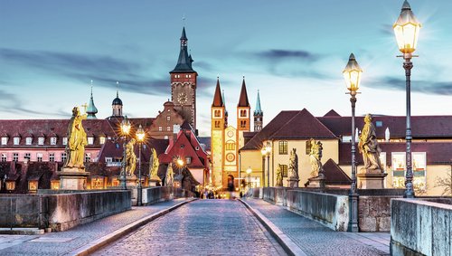 Die Alte Mainbrücke in Würzburg mit Statuen und Laternen bei Abendstimmung, im Hintergrund historische Gebäude und Türme unter blauem Himmel.