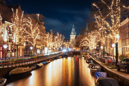 Blick auf eine weihnachtlich geschmückte und beleuchtete Gracht in Amsterdam mit mehreren Booten auf dem Wasser und historischen Gebäuden entlang des Kanals