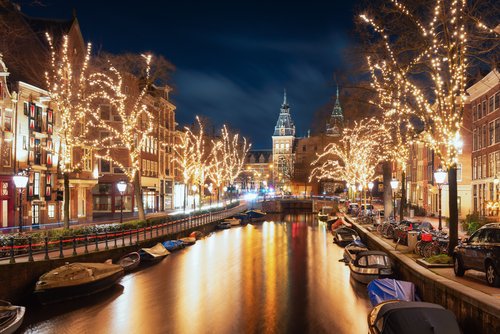 Blick auf eine weihnachtlich geschmückte und beleuchtete Gracht in Amsterdam mit mehreren Booten auf dem Wasser und historischen Gebäuden entlang des Kanals