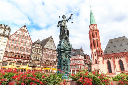 Statue der Justitia auf einem Brunnen in der Frankfurter Altstadt unter bewölktem Himmel. Blumen schmücken den Springbrunnen. Im Hintergrund historische Gebäude und Sonnenschirme.
