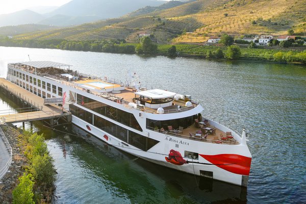 Flusskreuzfahrtschiff A-ROSA ALVA am Ufer eines Flusses mit Hügeln und Weinbergen im Hintergrund.