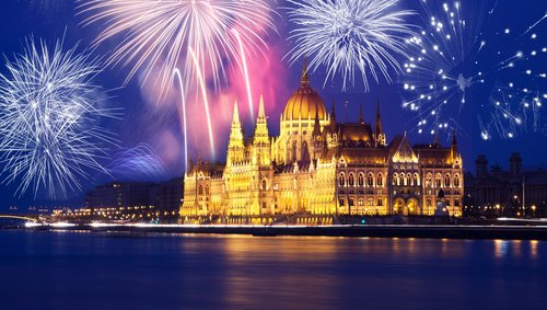 Blick auf das Parlamentsgebäude von Budapest bei Nacht. Es ist gold beleuchtet und über dem Gebäude ist ein Feuerwerk zu sehen.
