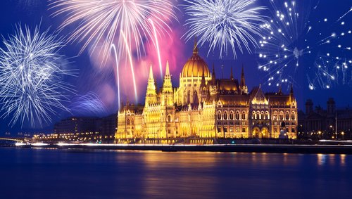 Blick auf das Parlamentsgebäude von Budapest bei Nacht. Es ist gold beleuchtet und über dem Gebäude ist ein Feuerwerk zu sehen.