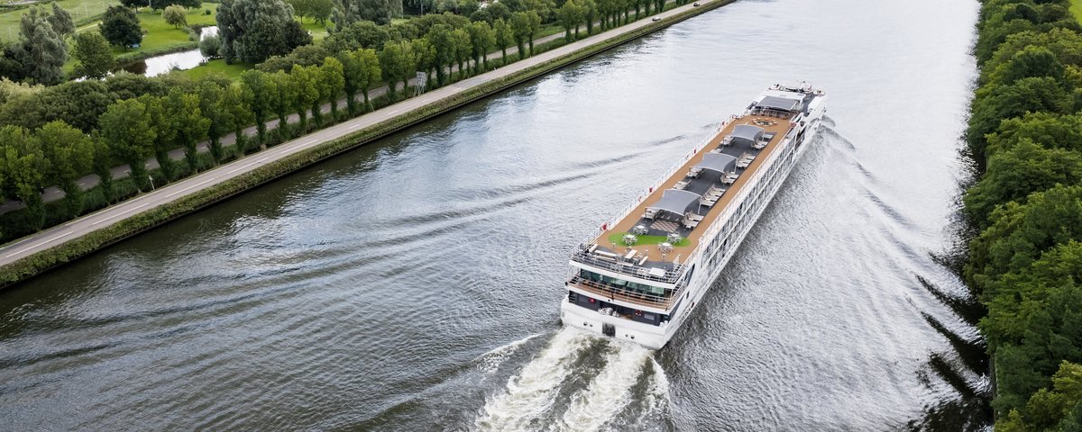 Ein Flusskreuzfahrtenschiff fährt auf dem Rhein am Ufer ein kleines Dorf, grüne Felder und Bäume. Das Sonnendeck ist mit Whirlpool, Sonnenliegen und Sonnensegeln ausgestattet.
