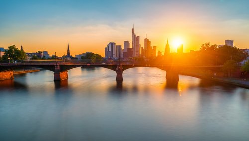 Frankfurter Skyline bei Sonnenuntergang, mit Wolkenkratzern und dem Main, welcher von einer Brücke überspannt wird, im Vordergrund,