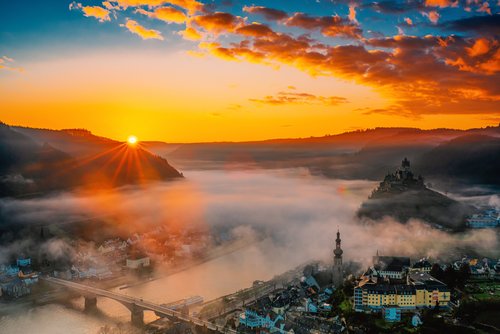 Blick auf die Stadt Cochem im Herbst, mit der Reichsburg auf einem Hügel, umgeben von Nebel und der Mosel im Vordergrund bei Sonnenuntergang