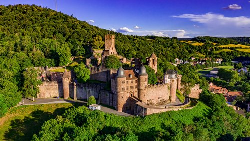Burg Wertheim thront bei strahlendem Sonnenschein auf einem Hügel, umgeben von grünen Bäumen, mit Blick auf die Stadt im Tal.