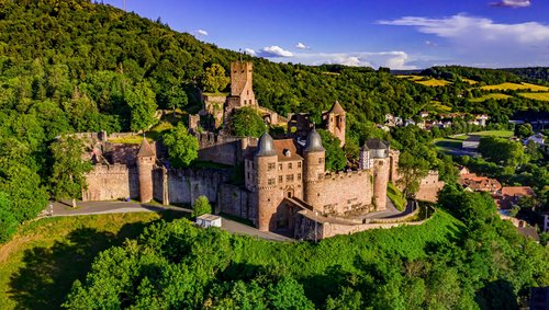 Burg Wertheim thront bei strahlendem Sonnenschein auf einem Hügel, umgeben von grünen Bäumen, mit Blick auf die Stadt im Tal.