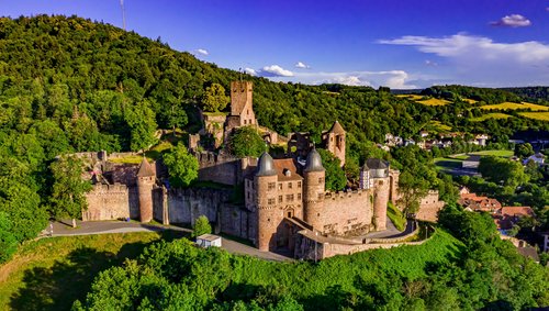 Burg Wertheim thront bei strahlendem Sonnenschein auf einem Hügel, umgeben von grünen Bäumen, mit Blick auf die Stadt im Tal.