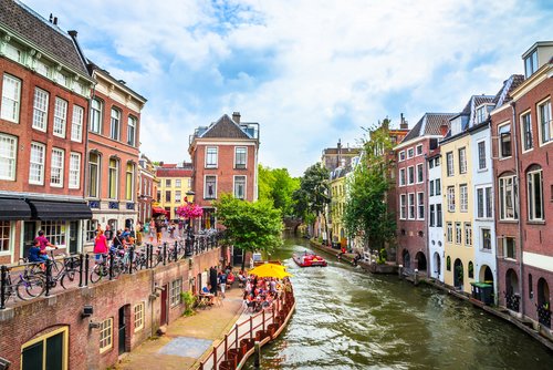 Blick auf einen belebten Kanal in Utrecht mit bunten historischen Gebäuden, Menschen am Ufer und einem Boot auf dem Wasser