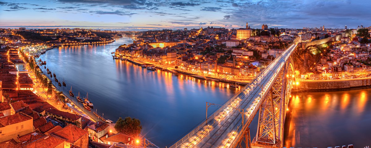 Blick auf die beleuchtete Dom-Luís-I-Brücke über den Douro-Fluss in Porto bei Abenddämmerung mit Stadtlichtern und historischen Gebäuden am Ufer