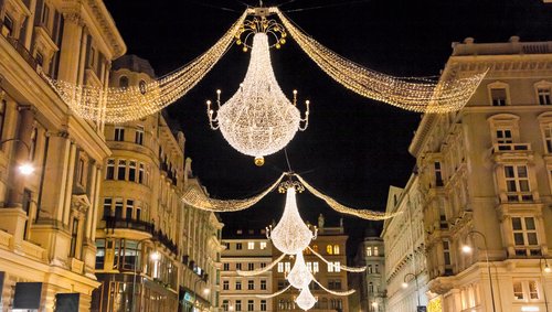 Weihnachtlich beleuchtete Einkaufsstraße in Wien bei Nacht, mit Lichterketen und Kronleuchtern, die zwischen die historischen Wohnhäuser gespannt sind.