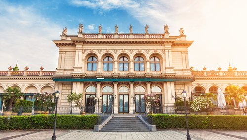 Beigefarbene Frontfassade eines barocken Gebäudes in Wien. Die Sonne strahlt golden über die Fassade hinweg.
