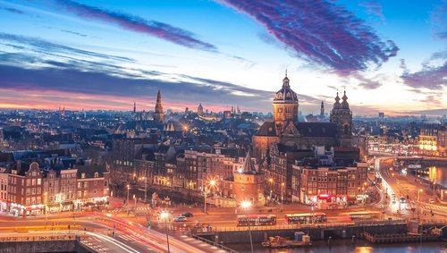 Abendliche Stadtansicht von Amsterdam mit historischen Gebäuden, beleuchteten Straßen und dramatischem Himmel mit Wolken