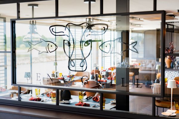 Blick durch eine Scheibe auf das Restaurant SENA GRILL im industriell-modernen Design auf der A-ROSA SENA bei Tageslicht mit gedeckten Tischen und bodentiefen Fenstern.