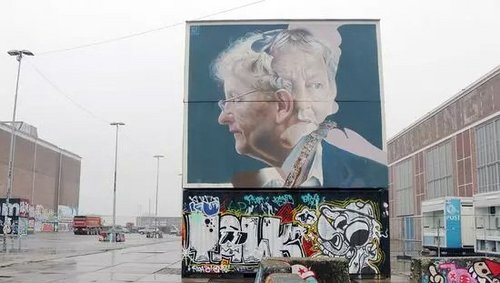 Großes Wandgemälde in Amsterdam zeigt das Porträt einer älteren Frau mit Brille und grauen Haaren, umgeben von bunter Graffiti-Kunst auf einer urbanen Betonfläche.