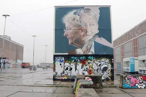 Großes Wandgemälde in Amsterdam zeigt das Porträt einer älteren Frau mit Brille und grauen Haaren, umgeben von bunter Graffiti-Kunst auf einer urbanen Betonfläche.