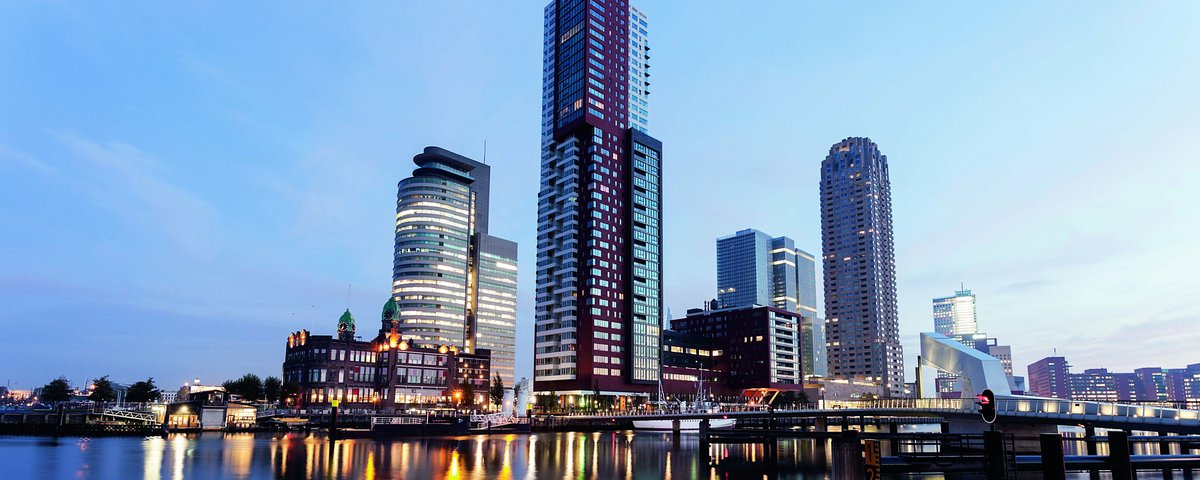 Skyline von Rotterdam an der Maas in der Abenddämmerung, mit modernen Hochhäusern an der Hafenkante. Im Fluss spiegeln sich die Lichter der Gebäude.