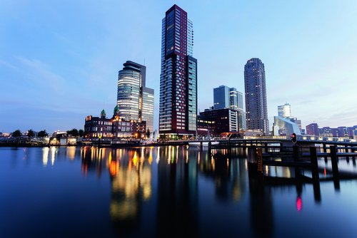 Skyline von Rotterdam an der Maas in der Abenddämmerung, mit modernen Hochhäusern an der Hafenkante. Im Fluss spiegeln sich die Lichter der Gebäude.