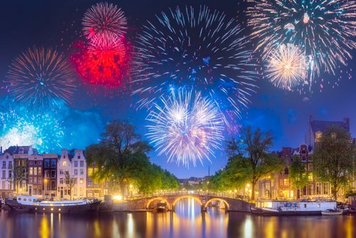 Feuerwerk über den beleuchteten Grachten vom Amsterdam bei Nacht. Am Kanal reihen sich historische Häuser und auf dem Wasser liegen Boote