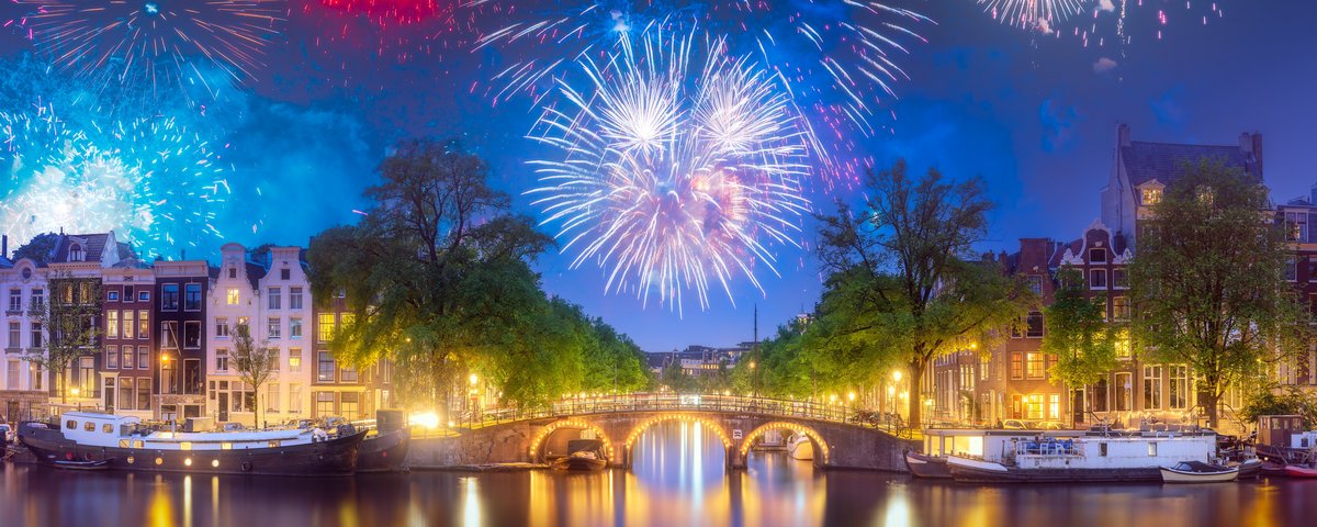 Feuerwerk über den beleuchteten Grachten vom Amsterdam bei Nacht. Am Kanal reihen sich historische Häuser und auf dem Wasser liegen Boote
