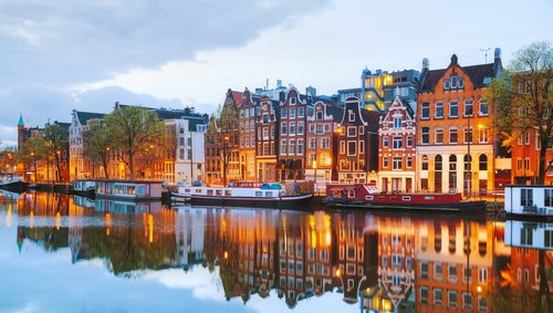 Blick auf Amsterdams Grachten mit historischen Gebäuden und Booten auf dem Wasser, während der Abenddämmerung.