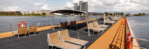 Das Sonnendeck mit zahlreichen Liegestühlen und Sonnensegeln bei Sonnenschein mit leicht bewölktem Himmel. Im Hintergrund Amsterdam. 
